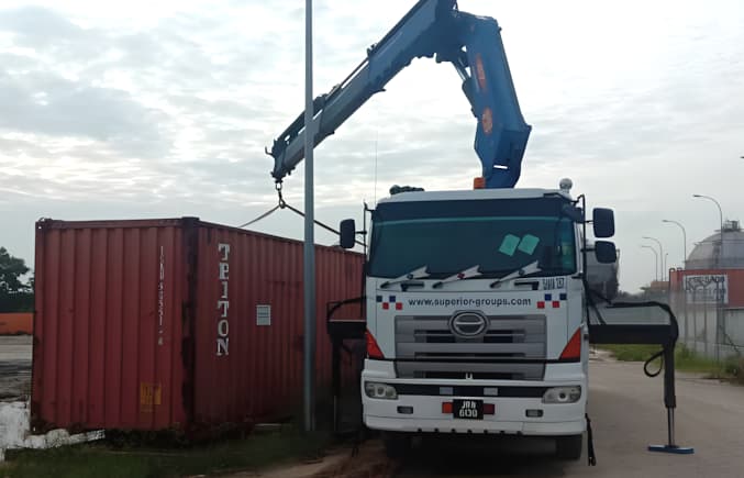 A lorry crane with its outriggers down, lifting a red shipping container.